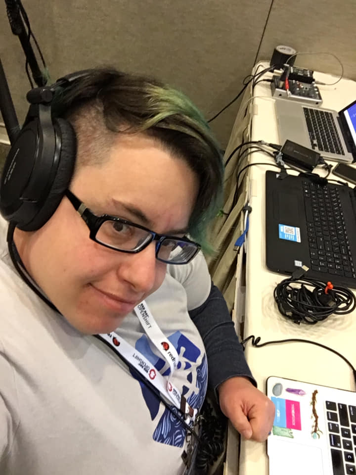 This is a photo of Rin at KubeCon. They are wearing a KubeCon shirt, and have headphones on. They have teal hair and wear glasses.